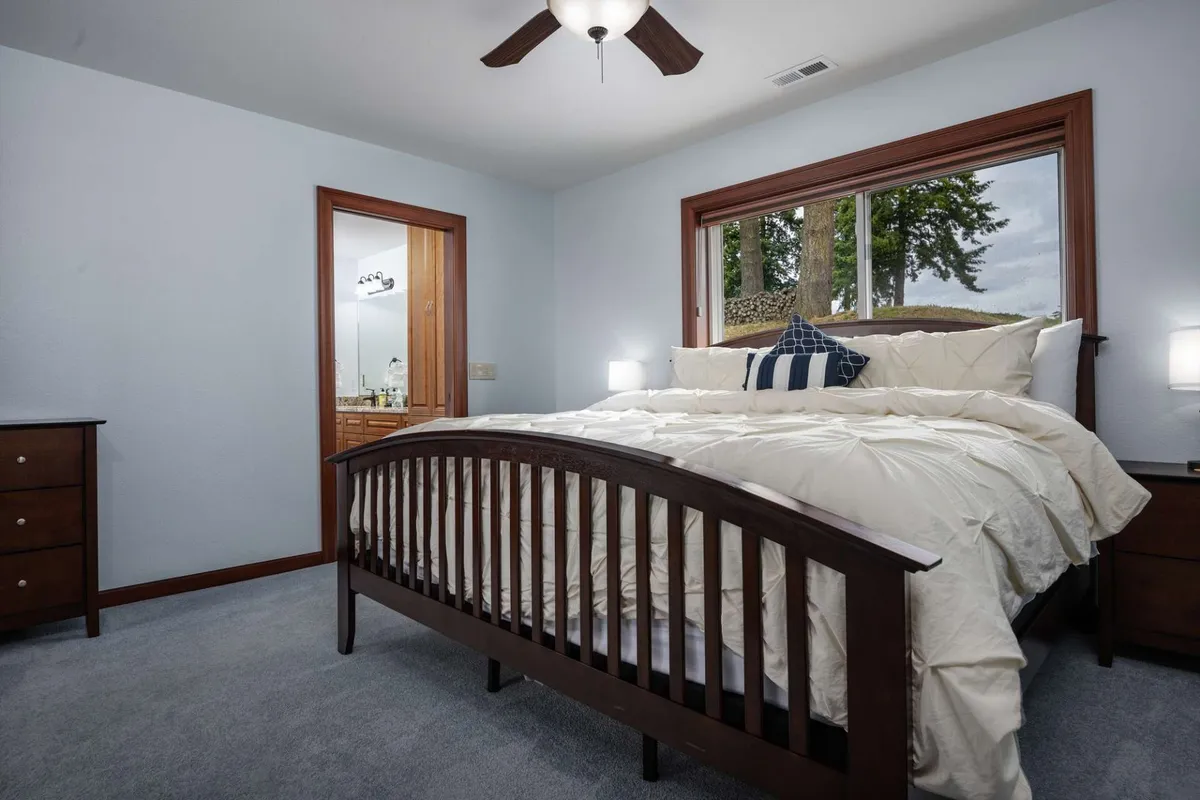A tranquil bedroom with a bed dressed in crisp white bedding, a large window offering views of the outdoors, and a nightstand with lamps for reading. The room is bathed in natural light, creating a peaceful environment ideal for unwinding.