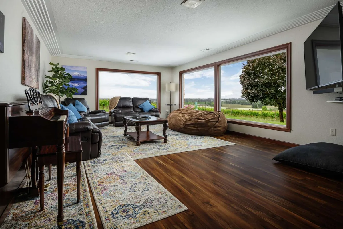 This image showcases a cozy, open-plan living room with large windows that offer panoramic views of the surrounding landscape. Comfortable leather seating is arranged for conversation and relaxation, complemented by a colorful area rug and a piano in the corner, inviting musical evenings. Sunlight floods the room, creating a bright and welcoming atmosphere.