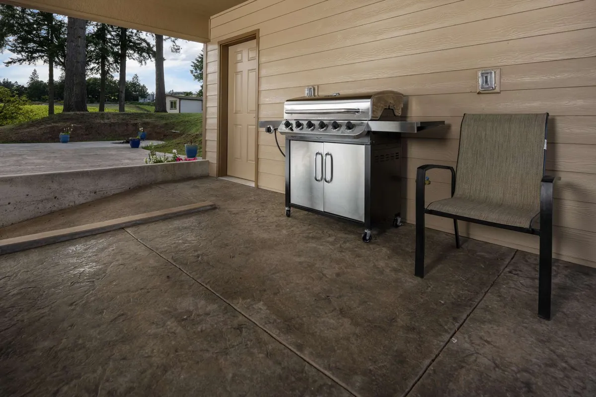 A BBQ grill is the focal point, accompanied by a comfortable chair and a door leading into the house. The setting is relaxed and casual, emphasizing outdoor cooking and enjoying meals outside in a homey atmosphere.