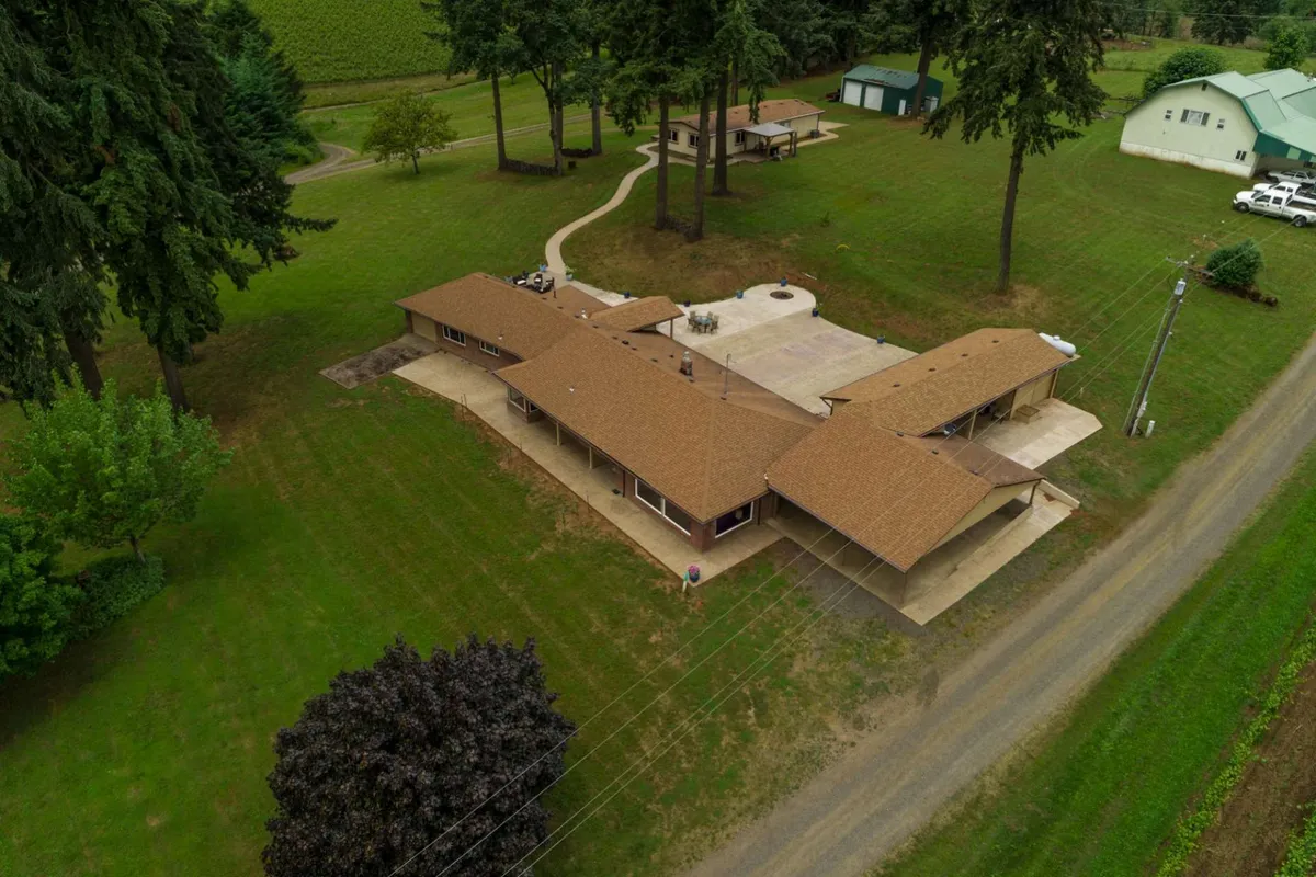 This image displays the main house with a spacious patio area, surrounded by lush trees and a welcoming walkway. The patio features outdoor seating ideal for gatherings and relaxation, all nestled in a peaceful natural setting.
