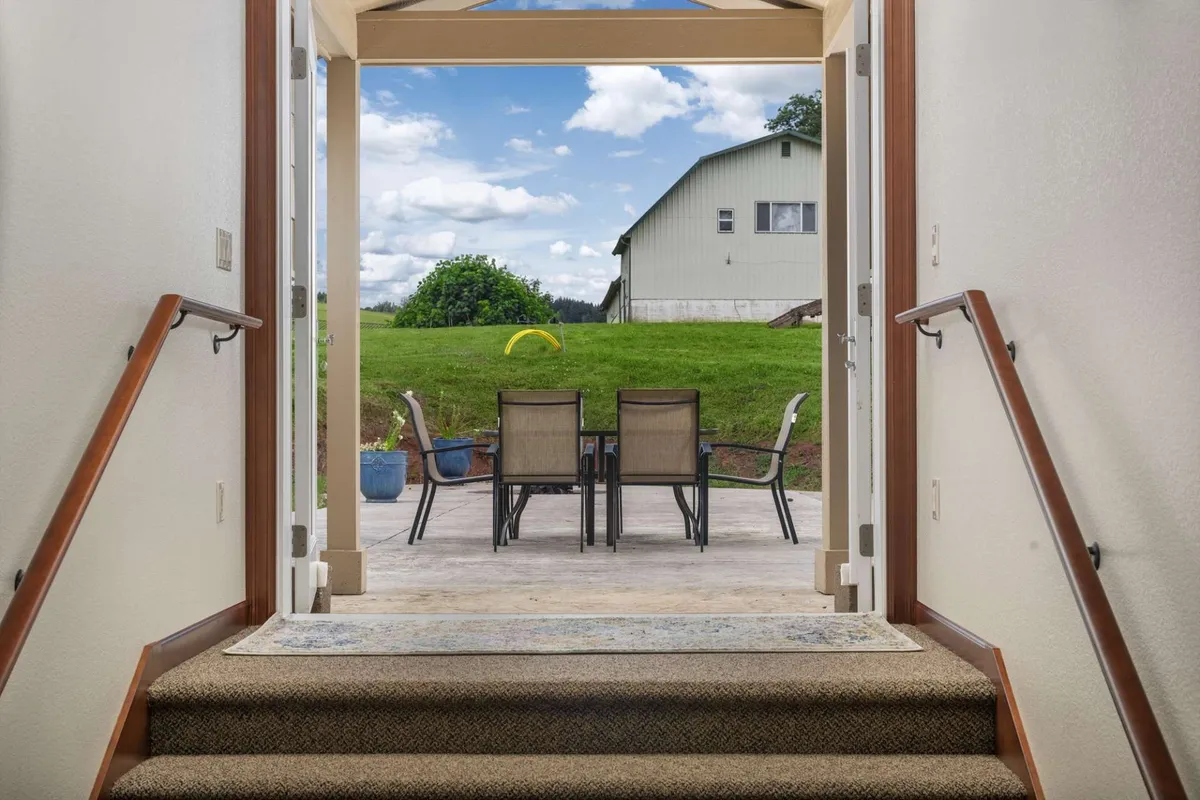 A staircase leads down to an outdoor seating area featuring comfortable chairs and a table, with a barn and open sky visible in the background. The patio is bathed in natural light, suggesting a peaceful spot for outdoor gatherings and relaxation, while connecting seamlessly to the surrounding landscape.