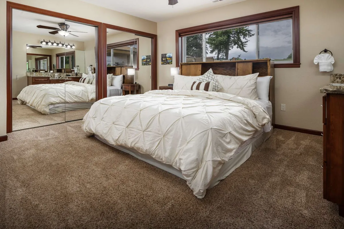 A serene bedroom featuring a neatly made bed with decorative pillows, mirrored closet doors, and wooden furniture. Natural light filters through the windows, creating a calm and inviting atmosphere ideal for relaxation.