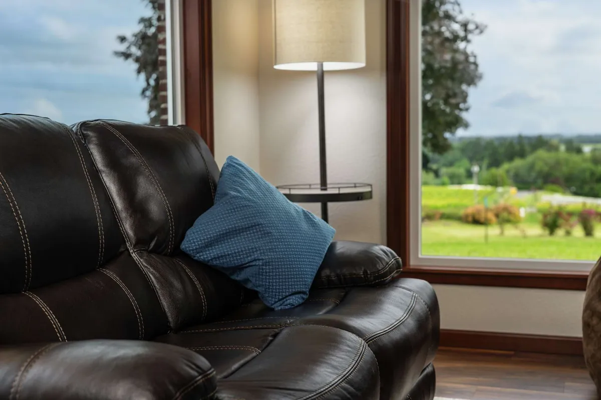 A spacious lounge with a black leather sofa, blue accent pillow, floor lamp, and a large window offering a view outside. The area feels inviting and bright, ideal for socializing, relaxing with a morning coffee, or enjoying a cozy evening indoors with friends or loved ones.